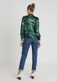 Green and black striped silk blouse with a high collar and long sleeves, paired with blue rolled cuff jeans and white block-heeled mules.