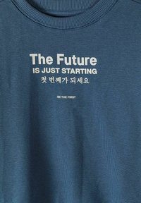Sweat-shirt bleu avec texte blanc : « L'avenir ne fait que commencer », caractères coréens et « Soyez les premiers » imprimés à l'avant.