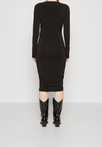 Černé šaty s dlouhým rukávem a přiléhavým střihem, s nařasenou texturou v oblasti pasu, v kombinaci s černými kozačkami po kolena a hladkým povrchem.