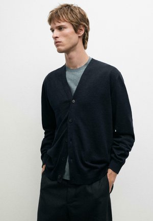 Ung mand iført en sort knapcardigan over en grå t-shirt, med hænderne i lommerne, stående foran en ensfarvet hvid baggrund.