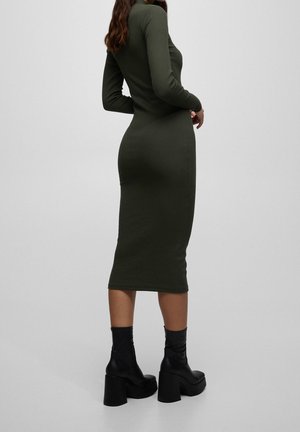 Femme portant une robe midi ajustée à manches longues de couleur vert olive et des bottines à plateforme noires, debout devant un fond uni.