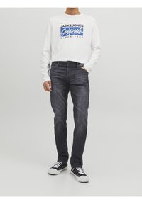 Mężczyzna w białej koszuli z długim rękawem Jack & Jones Originals, ciemnoszarych dżinsach i czarnych sneakersach z białymi sznurowadłami, stojący z jedną ręką w kieszeni.