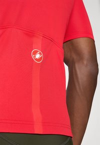 T-shirt de sport rouge à manches courtes en tissu texturé, doté d'une poche latérale et d'un logo circulaire blanc. Bande d'accent en rouge plus foncé.