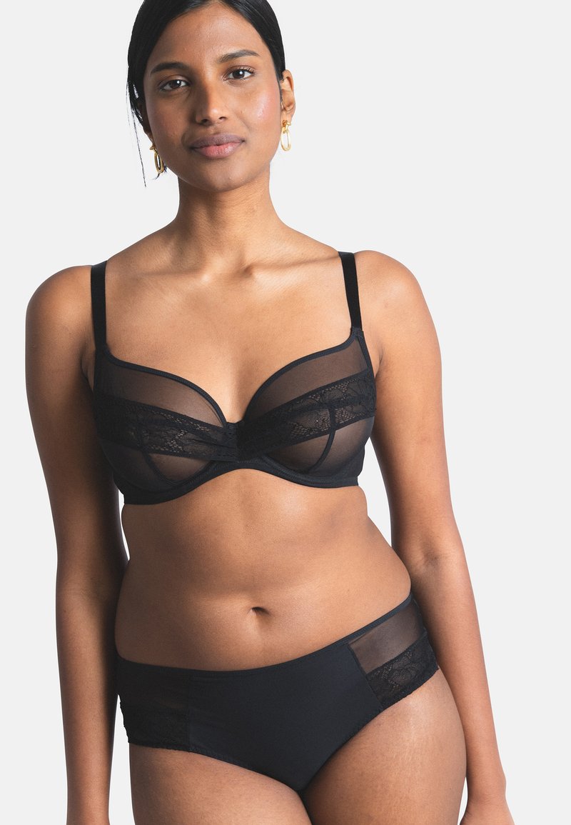 Frau trägt schwarzen durchsichtigen Spitzen-BH und passenden Slip, steht vor einfachem hellem Hintergrund mit neutralem Gesichtsausdruck.