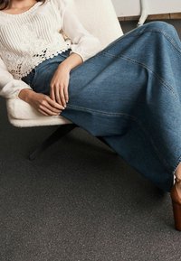 Denim maxi skirt with horizontal seams and a flared shape, paired with a white embroidered blouse. Brown wooden-heeled shoes.