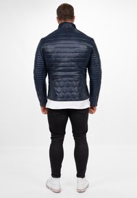 Marineblauwe leren jas met een gestructureerd hexagonaal patroon en horizontale gewatteerde strepen, voorzien van een hoge kraag en ritssluitingen aan de zijkanten. Zwarte jeans en witte sneakers.