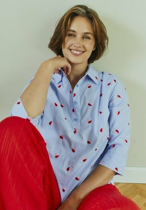 Vrouw zittend op de vloer, glimlachend, gekleed in een lichtblauw shirt met rode bladpatronen en felrode broek tegen een effen muur.