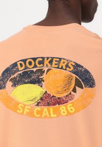Tricou de culoare piersică, cu un grafic uzat al fructelor și inscripția "Dockers SF CAL 86" în galben, având un accent rotund de culoare bleumarin.