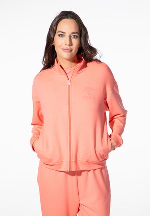 Frau mit langen dunklen Haaren, die einen korallenfarbenen Sweatshirt mit Reißverschluss und passende Hose trägt, mit den Händen in den Taschen, stehend vor einem weißen Hintergrund.