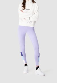 Leggings viola con un design a blocchi di colore e dettagli blu, abbinati a una felpa bianca con zip e sneakers bianche.