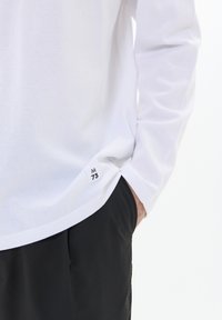 Witte langemouwen shirt met afgeronde zoom en zijkleed met de tekst "M 73". Gedragen over zwarte broek, met een soepele katoentextuur.