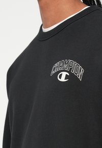 Sweatshirt preto feito de algodão, com decote redondo e logótipo branco "CHAMPION" acima de um "C" estilizado no peito.