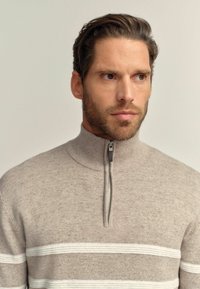 Khaki-Zipfelpullover mit hohem Kragen, ausgestattet mit weißen horizontalen Streifen und einer strukturierten Oberfläche. Metalldetail mit Reißverschluss am Hals.