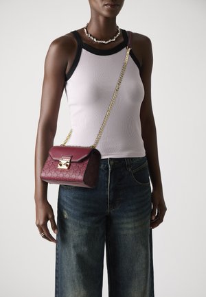 Burgundy läder crossbody-väska med guldbeslag, strukturerat mönster och kedjesnurra, bärs tillsammans med en åtsittande grå linne och mörka jeans.