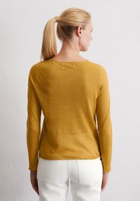 Langärmliger, senffarbener Pullover aus leichtem, strukturiertem Stoff; verfügt über einen runden Halsausschnitt und einen leicht durchsichtigen Finish. Getragen mit weißen Hosen.