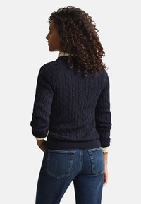 Pull en maille torsadée bleu marine avec manches longues, poignets et ourlet côtelés. Porté sur une chemise claire à col, associé à un jean bleu ajusté.