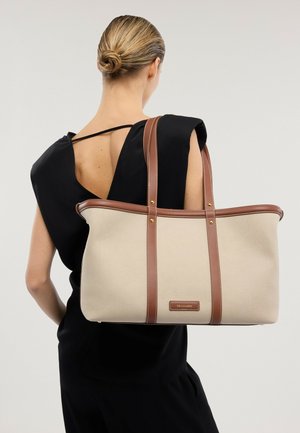 Mujer con cabello rubio recogido en un moño bajo, vistiendo un vestido negro con la espalda descubierta, llevando un gran bolso tote beige con tiras de cuero marrón.