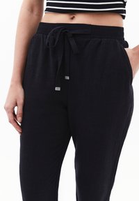Schwarze, leichte, atmungsaktive Hosen mit einem elastischen Bund, einer Kordel und seitlichen Taschen. Glatte Textur, lässiges Design.