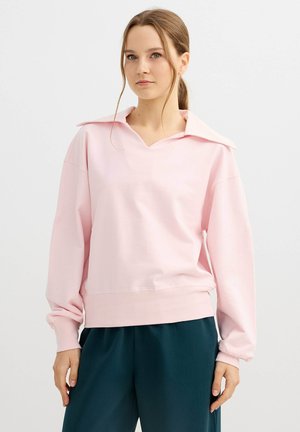 Joven mujer con el pelo recogido, vistiendo un top de manga larga rosa claro con un cuello ancho y pantalones verde oscuro, de pie frente a un fondo liso.