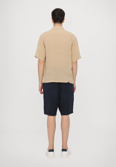 Beige short-sleeve shirt with a textured finish, paired with dark navy shorts. White sneakers with green accents complete the outfit.