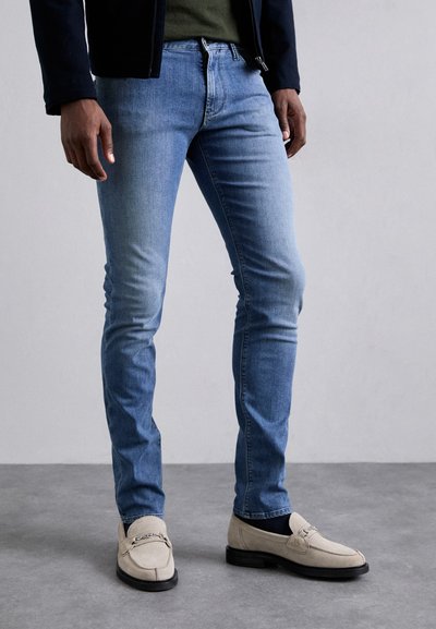 Jasnoniebieskie dżinsy o slim fit, charakteryzujące się gładką teksturą i przeszyciami. Połączone z beżowymi mokasynami.
