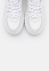 Bílé kožené tenisky s bílými tkaničkami zobrazené shora na jednobarevném bílém pozadí.