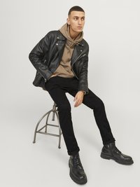 Un jeune homme assis sur un tabouret en métal, portant une veste en cuir noire, un sweat à capuche brun, un pantalon noir et des bottes noires, regardant sur le côté.