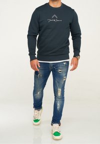 Marinblå sweatshirt med rund halsringning, elastiska mudd och nederkant. Kombinerad med slitna blå jeans och vita sneakers med gröna detaljer.