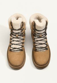 Smeđe nubuk planinarske čizme s kremastim umetkom od umjetnog ovčjeg krzna, imaju tamne vezice i metalne rupice za sigurno pričvršćivanje te robusnu potplat.