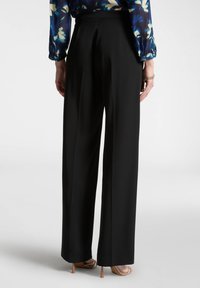 Pantaloni neri a gamba larga con una texture liscia, dotati di vita alta e pieghe ben definite, abbinati a tacchi nudi con cinturini.