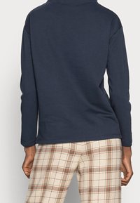 Maglietta a maniche lunghe blu navy in tessuto morbido, con colletto alto e vestibilità rilassata, abbinata a pantaloni a quadri crema con accenti arancioni.