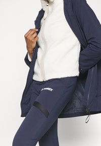 Mörkblå zip-jacka över ett vitt fleece-lager, tillsammans med mörkblå leggings med en svart rand och logotyp. Mjuka texturer, modern design.