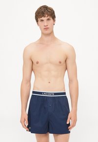 Bikini navy cu țesătură ușoară, talie elastică, cu un logo Lacoste de culoare albastru deschis și închidere cu un buton în față.