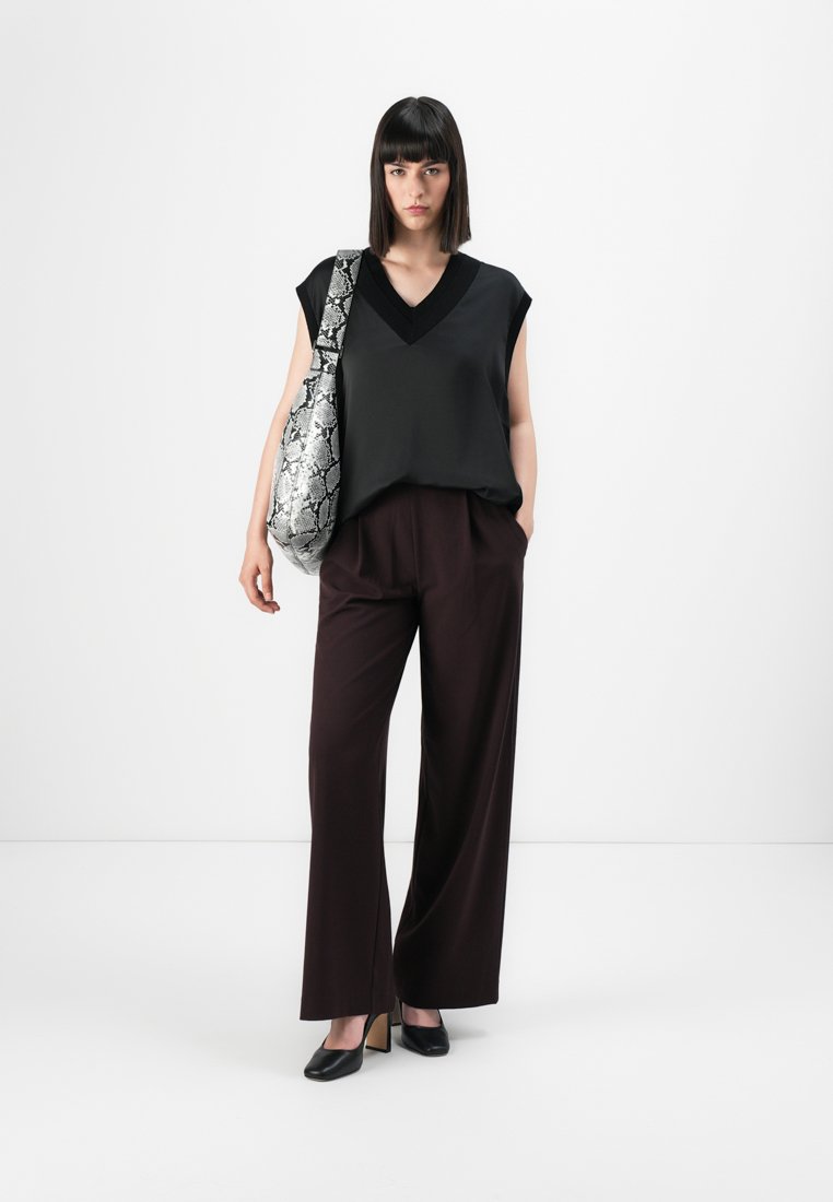 Femme portant un top sans manches noir à col en V, un pantalon large sombre, des talons noirs et portant un grand sac à épaule en simili-cuir argenté à motifs serpent.