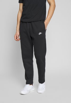 Person iført sorte Nike joggingbukser, sort skjorte og hvide sneakers, stående med den ene hånd i lommen mod en ensfarvet baggrund.