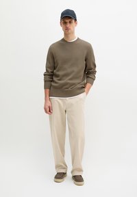 Homme portant un pull côtelé marron, un pantalon beige, des chaussures marron sans lacets et une casquette sombre, debout avec une main dans la poche devant un fond blanc.