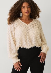 Vrouw met lang krullend haar, gekleed in een crèmekleurige, textuur V-halstrui, zwarte broek en hangende rhinestone oorbellen, staand tegen een effen achtergrond.