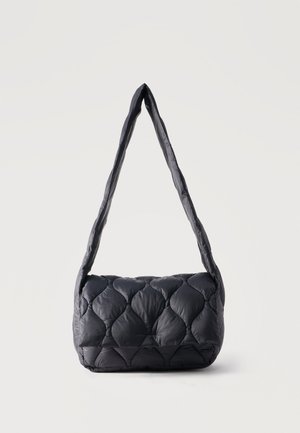 Μαύρη καπιτονέ τσάντα ώμου με επενδεδυμένη υφή και μακρύ, απαλό λουρί, τοποθετημένη σε απλό ανοιχτόχρωμο φόντο.