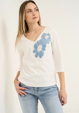 Frau in weißem V-Ausschnitt-Shirt mit blauer Blumenapplikation, Dreiviertelärmeln und hellblauen Jeans, stehend vor einem schlichten hellen Hintergrund.