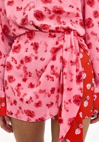 Blouse fleurie rose avec des motifs de roses rouges, manches longues et taille nouée, associée à une jupe rouge ornée de petits accents floraux roses.