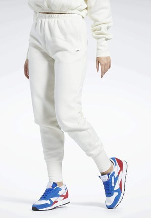 Personne portant un pantalon de jogging blanc et des baskets bleues, blanches et rouges, debout avec une jambe légèrement pliée sur un fond blanc.