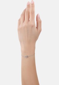 Mano con unghie smaltate di rosa chiaro che indossa un delicato braccialetto d'argento con due anelli intrecciati sul polso su sfondo bianco.