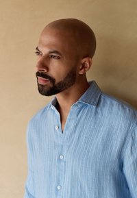 Chemise en lin rayée bleu clair avec un col officier, dotée de boutons blancs et d'une coupe décontractée. Texture lisse et détails minimaux.