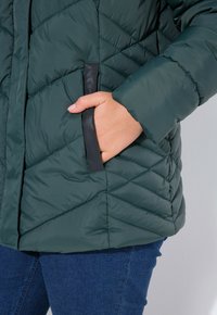 Grøn quiltet pufferjakke med diagonale syninger, sorte lommenkanter og en afslappet pasform, parret med mørkeblå jeans.