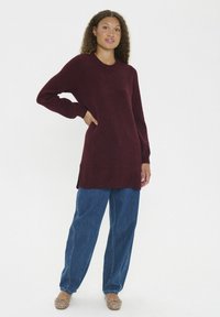 Vrouw met krullend haar, gekleed in een lange bordeauxrode trui, losse blauwe jeans en beige gestreepte ballerina's, staande met één hand op haar heup.