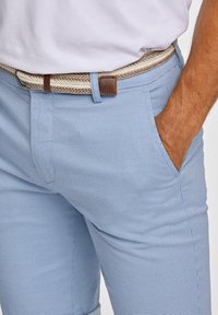 Ljust blå texturerade byxor med sidofickor, kompletterade av ett vävt beige och brunt bälte. Tyget har ett subtilt mönster.