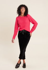 Rosa Strickpullover mit strukturiertem Muster, kombiniert mit schwarzen Cordhosen mit einem Kordelzugbund und umgeschlagenen Säumen. Schwarze Loafer runden den Look ab.