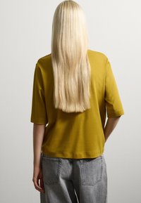 Top giallo a maniche corte con una vestibilità comoda, realizzato in tessuto morbido. Abbinato a jeans grigi chiari a gamba larga, visto da dietro.