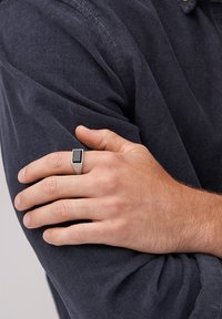 Bague rectangulaire en argent avec un centre noir, portée sur une main à la peau claire, reposant sur une manche en tissu sombre.