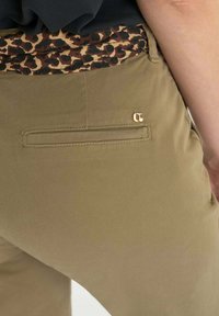 Pantalons khaki avec une texture lisse, dotés d'une poche arrière avec une couture horizontale et un logo en ton doré au-dessus. Ceinture à motif léopard visible.